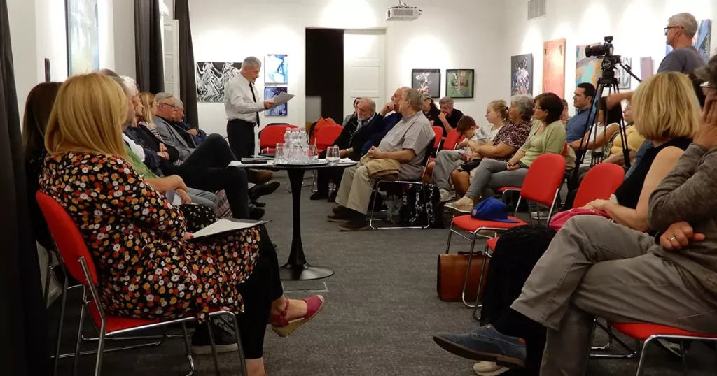Ungarndeutsche Kunst und Literatur in Fünfkirchen VUdAK-Vernissage im Lenau Haus Von Gabriella Sós Ein besonderes Kulturerlebnis bot den Interessenten der Verband ungarndeutscher Autoren und Künstler (VUdAK), der am 26. September 2025 zur Vernissage seiner neuen temporären Ausstellung mit dem Titel „Quadriga“ mit anschließender Lesung ungarndeutscher Gegenwartsautoren nach Fünfkirchen einlud. In seiner Eröffnungsrede informierte 1. Vorsitzender Johann Schuth über die vielfältigen Tätigkeiten von VUdAK, beginnend mit der Vorstellung einer thematischen Rollup-Ausstellung, die mittlerweile schon Ungarn bereist und wöchentlich in einer anderen Bildungseirichtung ausgestellt die Arbeit des Verbandes den Schülern näher bringt. Als Begleitprogramm finden dazu auch immer Lesungen mit den Autoren der Literatursektion statt. In den Schulen verteilt der Verband außerdem reichlich Informationsmaterial über ungarndeutsche Künstler und Autoren, welches als Unterrichtsstoff eingesetzt werden kann. Jährlich veranstalten sie außerdem mit dem Titel VUdAK-Gespräche dreitägige thematische Aufenthalte landesweit an unterschiedlichen Orten, wo der Verein zusammenkommt, ausstellt und sich auch mit dem Publikum persönlich trifft − auch die Ausstellungseröffnung in Fünfkirchen war Teil dieser Programmreihe. Im Veranstaltungssaal des Lenau Hauses sind diesmal Werke von vier Mitgliedern der Künstlersektion von VUdAK, Bernadett Breszkovics, Erzsébet Horváth, Erzsébet Lieber und Zsuzsa Trieb zu betrachten. Von den Malerinnen waren bei der Vernissage die ersten zwei persönlich anwesend. Durch die aktuelle Ausstellung führte ein, wie Vorsitzender Johann Schuth betonte, hochgeschätztes Mitglied des Verbandes, Kunstsachverständiger Johann Wolfart jun., der regelmäßig Rezensionen über Werke und Ausstellungen von VUdAK veröffentlicht und hauptberuflich im professionellen Kunsthandel tätig ist. Wolfart entschied sich als Auftakt für ein Gespräch mit den Künstlerinnen Breszkovics und Horváth und leitete das Gespräch stilvoll – da die Veranstaltung ja im Lenau Haus stattfand – mit einem Zitat aus Nikolaus Lenaus Faust ein: „Behaupten will ich fest mein starres Ich /Mir selbst genug und unerschütterlich, /Niemandem hörig mehr und untertan, /Verfolg ich in mich einwärts meine Bahn.“ Auf die Kunst übertragen interpretierte Wolfart dieses Zitat als Verbildlichung der künstlerischen Freiheit, die die bildenden Künstler verfolgen. Erzsébet Horváth ist laut Wolfart eine vielseitige Künstlerin, die mit unterschiedlichen Techniken arbeitet. In ihrem Repertoire befinden sich Installationen, Objekte und Gemälden sowie auch Plastiken. Ein Kunststipendium und vierjähriger Aufenthalt in München wirkten besonders inspirierend auf Horváth, sie begann dort mit Kunststoff zu arbeiten und durch die dortige Papierwerkstatt kam sie auch mit selbstgeschöpftem Papier und deren Verarbeitungsmöglichkeiten in Kontakt. Ihre Kunst zeichnet sich vor allem vom Spiel von Licht und Schatten aus, gerne verwendet ganz alltägliche Materialien, wie Folien, Papier und Farben. In der Quadriga-Ausstellung sind von ihr fünf Arbeiten zu besichtigen. Das Subjektive, Erinnerungen und innere Bilder und deren Verhältnis zur Außenwelt bilden die wichtigsten Inhalte und gleichzeitig Inspiration der Arbeiten von Bernadett Breszkovics. In ihren recht farbenfrohen Gemälden, wie auch in ihren modernen Installationen verwendet sie oft Textilien in Kombination mit Farben und bringt diese durch die Schichtung der einzelnen Texturen zur Geltung. Licht und Schatten sind auch in ihrer Arbeit ständig wiederkehrende Motive. Als Künstlerin möchte sie, wie sie beim Gespräch betonte, auf die Vielschichtigkeit der Visualität reflektieren. Bei der Ausstellung sind von ihr ebenfalls fünf Arbeiten zu sehen. Kunst und Literatur gehen Hand in Hand bei VUdAK. Im Anschluss an die Ausstellungseröffnung fanden Lesungen aus den Werken von Christina Arnold (Nadasch), Susi Szabó (Surgetin), Robert Becker (Surgetin), Koloman Brenner (Ödenburg), Alfred Manz (Baja), Peter Wess (Feked), Diana Feuerbach (Leipzig), Josef Michaelis (Schomberg) und Stefan Valentin (Schorokschar) von der VUdAK-Literatursektion statt. Erfrischende Neuigkeiten präsentierten die Gegenwartsautoren in den unterschiedlichsten Genres von Gedichten bis hin zu Auszügen aus Novellen, im Dialekt und in der Hochsprache. Besonders realitätsnah waren die Auseinandersetzungen mit existentiellen Themen unserer Gegenwart, wie den Generationsunterschieden, dem medialen Körperkult, der Künstlichen Intelligenz und dem technischen Fortschritt, aber auch etwas ernstere Themen, wie Überlegungen zu Spracherhalt und Sprachverlust, Identität und Identitätsverlust wurden behandelt, außerdem fanden auch internationale Kriminalität, Multikulturalität und durch Novellenauszüge Einblicke in das moderne Familienleben und in den Schulalltag ihren Platz in diesem prickelnden, köstlichen Cocktail der ungarndeutschen Gegenwartsliteratur, durch hervorragende Darbietungen der Autoren. Fotos: Lenau Haus Fünfkirchen Keretezett, külön kiemelt szöveg: Die VUdAK-Ausstellung Quadriga kann bis Ende Oktober 2025 im Veranstaltungssaal des Lenau Hauses (Fünfkirchen, Munkácsy M. Str. 8) und im Anschluss ab November im Otto-Heinek-Saal im Haus der Ungarndeutschen (Budapest, Lendvay Str. 22) besichtigt werden. Weitere Informationen